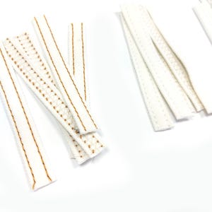 Può includere: Diverse strisce di tessuto bianco con cuciture e motivi decorativi marroni sono disposte su una superficie bianca. Le strisce variano nel design, alcune con un motivo a pois, altre con cuciture lineari.