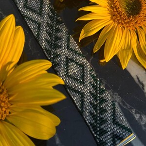 May include: A beaded bracelet with a geometric pattern in shades of gray and green, set against a dark background with bright yellow sunflowers. The bracelet has silver-toned metal ends and a clasp.