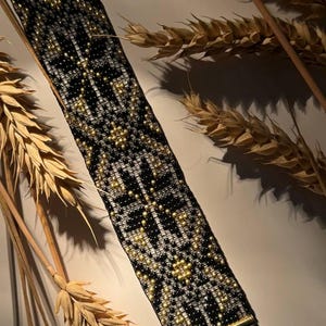 Puede incluir: Una pulsera de cuentas con un patrón geométrico negro, blanco y dorado. La pulsera tiene un cierre dorado y está decorada con pequeñas cuentas redondas. El fondo incluye espigas de trigo.