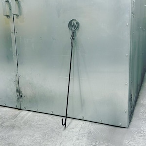 May include: A black metal fireplace poker with a round handle featuring the letter "W". The poker is leaning against a silver metal wall.