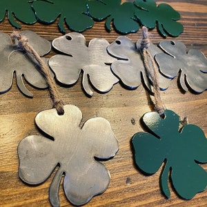May include: A set of four-leaf clover ornaments, two are green and two are silver. The ornaments are made of metal and have a rustic look. They are hanging from twine.
