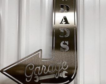 May include: A metal sign with an arrow pointing left. The sign says "Garage" with a wrench symbol and "Dad's" in a banner above the arrow.