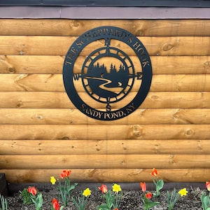 Puede incluir: Un letrero de metal negro con un diseño de brújula y el texto "The Sheppard's Hook Sandy Pond, NY". El letrero está montado en una pared de madera con un jardín de flores debajo.