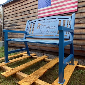 Puede incluir: Un banco de metal azul con una inscripci&oacute;n personalizada que dice "En memoria cari&ntilde;osa de Natalie Bauer "Vive a tope"". El banco est&aacute; sentado en una plataforma de madera frente a un edificio de madera con una bandera estadounidense colgada en la pared.