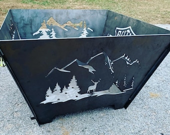 May include: A black metal fire pit with a cut-out design of mountains, trees, and a deer. The fire pit has a square shape with rounded corners and a slightly angled design. The fire pit is sitting on a gravel surface.