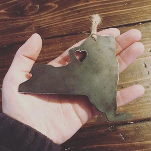 May include: A metal ornament shaped like the state of New York with a heart cut out in the center. The ornament is a rustic brown color and has a twine hanger.