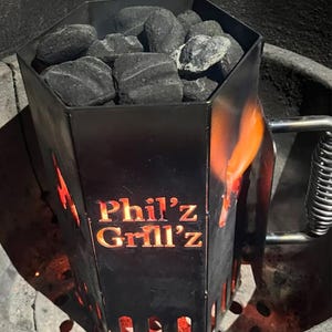 May include: A black metal charcoal chimney starter with a hexagonal shape. The chimney is filled with charcoal briquettes and is lit with flames visible through the openings. The chimney has the text "Phil's Grill'z" printed on the side.