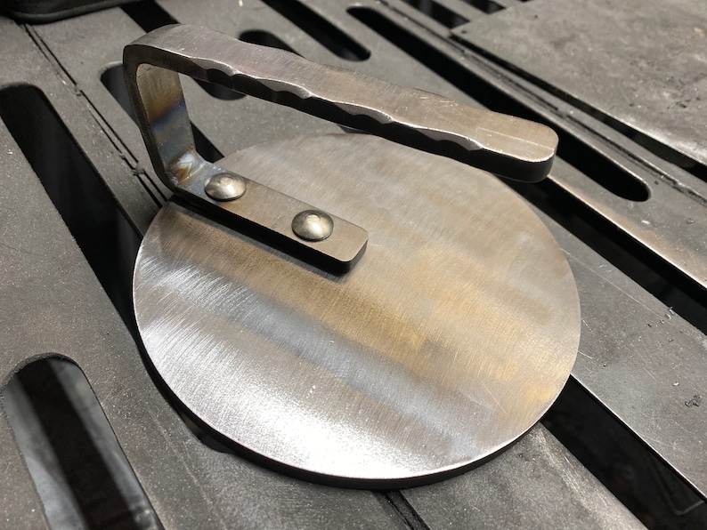 May include: A round, metal pizza stone with a handle. The stone has a brushed finish and is made of durable metal.