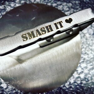May include: A silver metal tool with a handle that says "SMASH IT" with a heart symbol. The tool is on a silver metal surface.