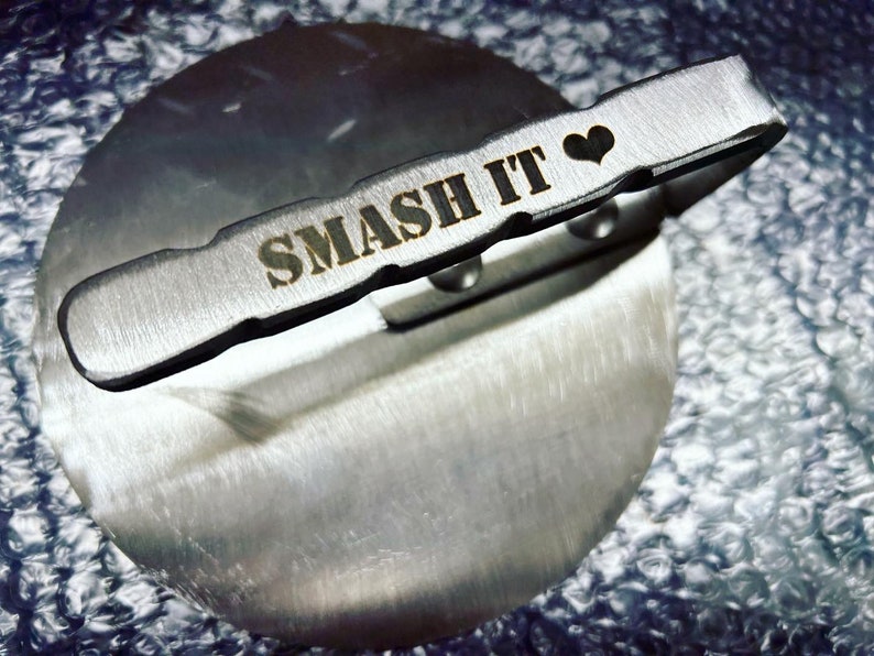 May include: A silver metal tool with a handle that says "SMASH IT" with a heart symbol. The tool is on a silver metal surface.