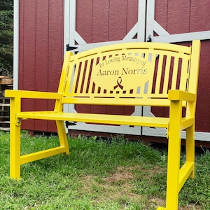 Puede incluir: Un banco de metal amarillo con una cinta negra y la inscripci&oacute;n "In Loving Memory of Aaron Norris".