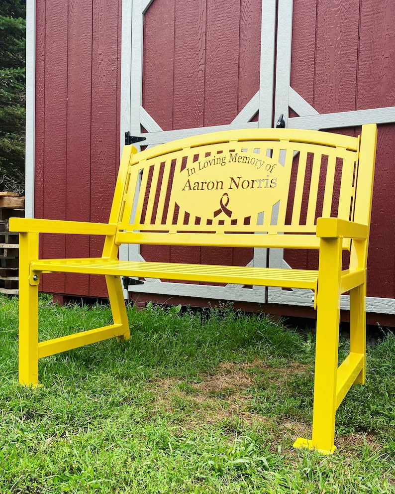 Puede incluir: Un banco de metal amarillo con una cinta negra y la inscripci&oacute;n "In Loving Memory of Aaron Norris".