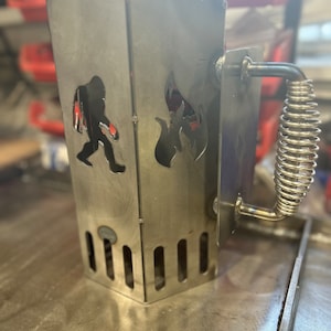 May include: A silver metal charcoal chimney starter with a spring-loaded handle. The chimney has a cutout of a fire and a silhouette of a Bigfoot.