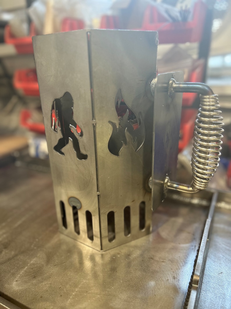 May include: A silver metal charcoal chimney starter with a spring-loaded handle. The chimney has a cutout of a fire and a silhouette of a Bigfoot.