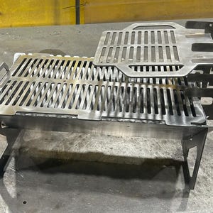 May include: A stainless steel portable grill with a rectangular design. The grill features a slotted cooking surface and a removable top grate. The legs are angled for stability, and the overall design is industrial.