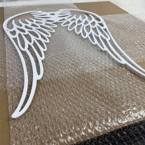 May include: White painted wooden angel wings with a detailed design. The wings are laid out on a sheet of bubble wrap.