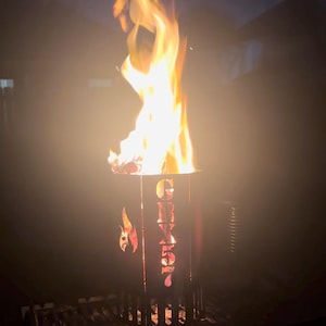 May include: A black metal fire pit with the word "Grizzly" cut out of the side. The fire pit is on a black metal grate and has a fire burning inside.