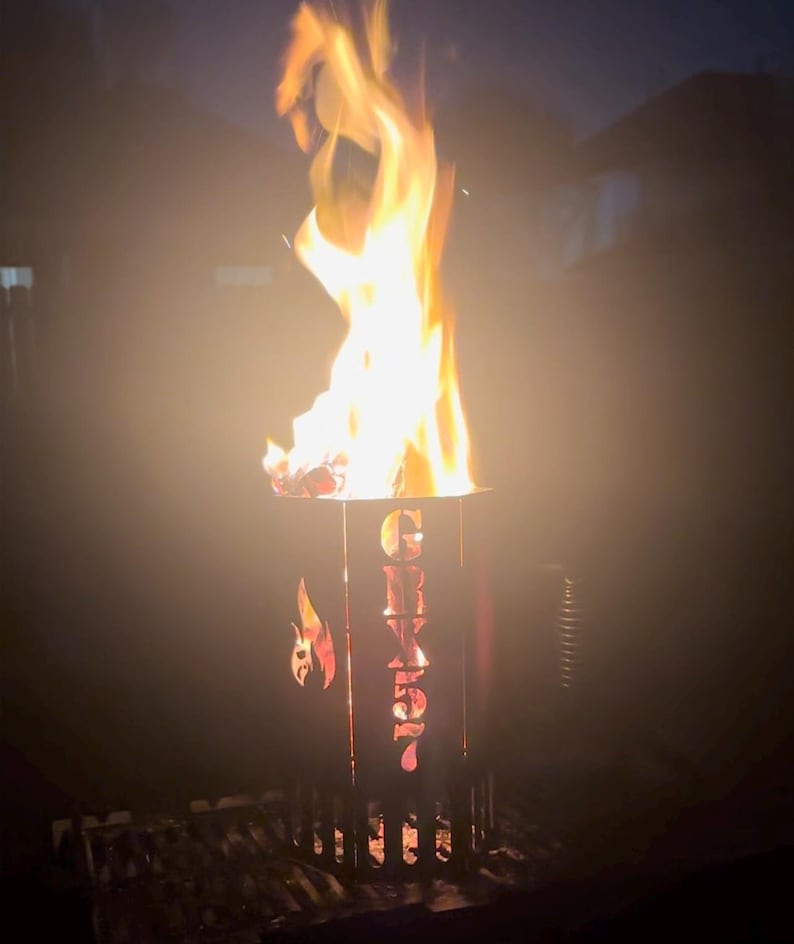 May include: A black metal fire pit with the word "Grizzly" cut out of the side. The fire pit is on a black metal grate and has a fire burning inside.