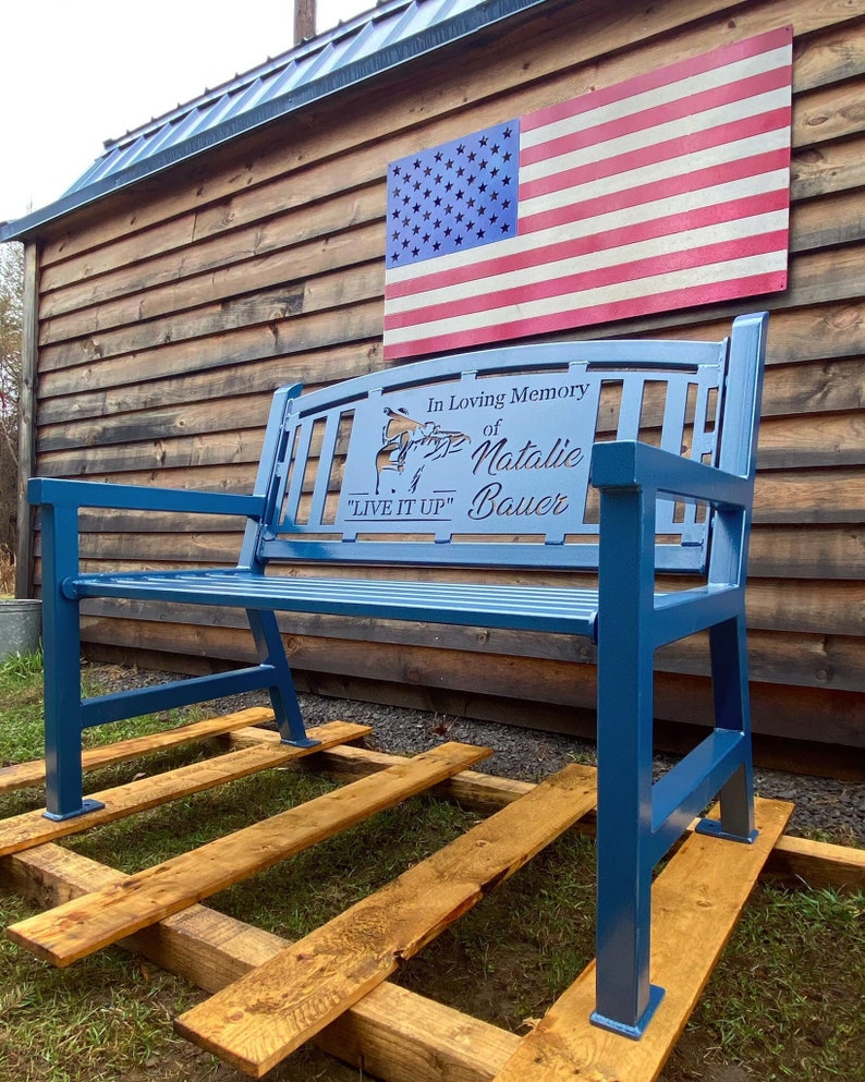 Puede incluir: Un banco de metal azul con una inscripci&oacute;n personalizada que dice "En memoria cari&ntilde;osa de Natalie Bauer "Vive a tope"". El banco est&aacute; sentado en una plataforma de madera frente a un edificio de madera con una bandera estadounidense colgada en la pared.