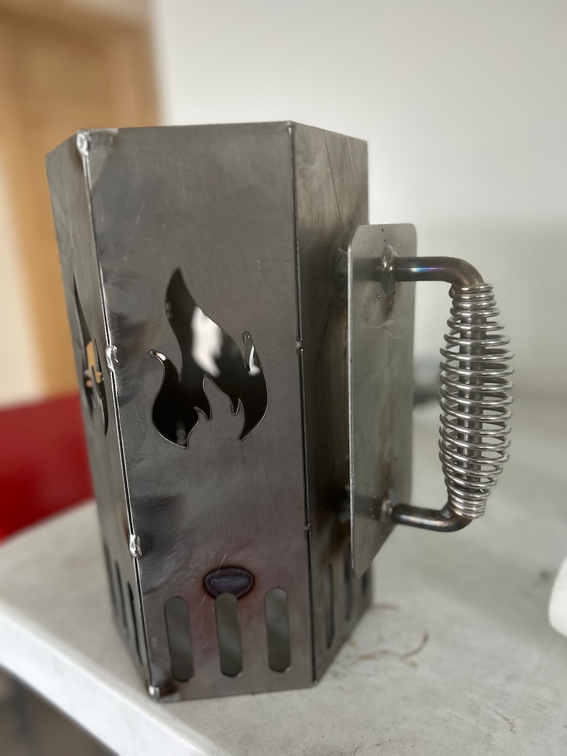 May include: A metal charcoal chimney starter with a spring-loaded handle and a flame cutout design.