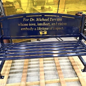 Puede incluir: Un banco de metal azul con una inscripci&oacute;n dorada que dice "Para el Dr. Michael Terrnini cuyo amor, intelecto y visi&oacute;n encarnan una vida de impacto". La inscripci&oacute;n est&aacute; en la parte posterior del banco e incluye dos huellas de manos.