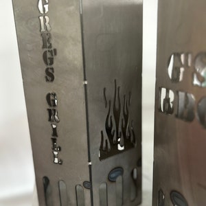 May include: Two metal fire pit stands with cut-out flames and the words "Greg's Grill" and "G's BBQ" on the front.