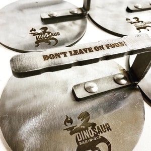 May include: A set of six round metal pizza peels with a handle. The peels are engraved with the words "Dinosaur Bar-B-Que" and "Don't Leave On Food!"