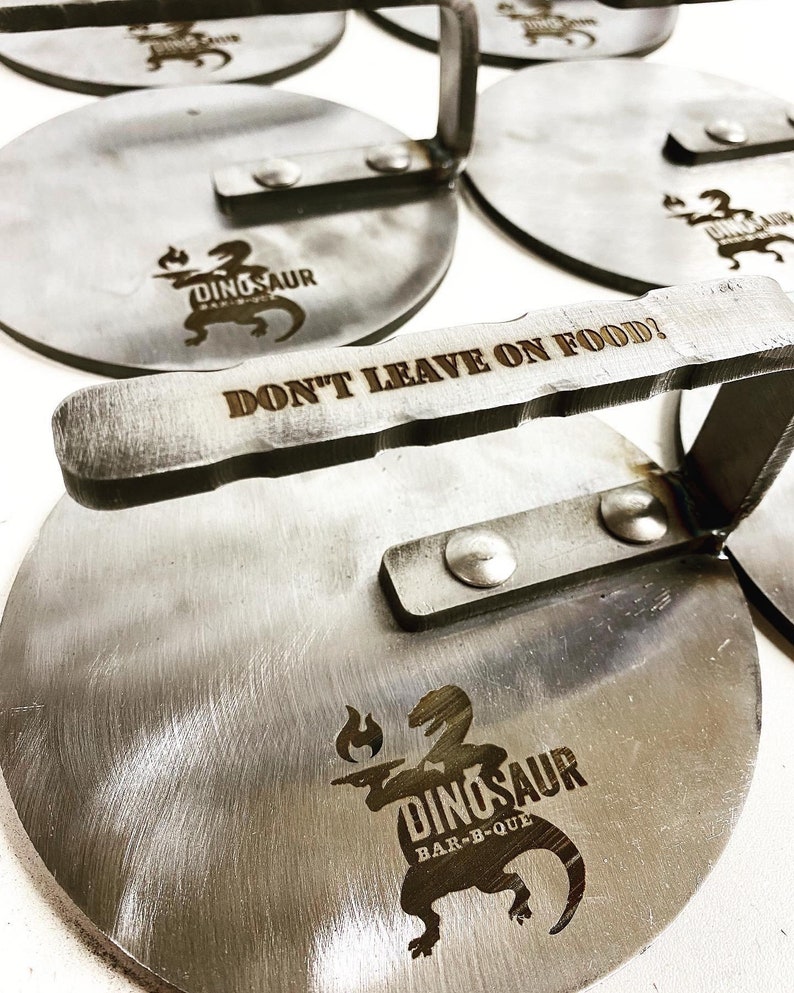 May include: A set of six round metal pizza peels with a handle. The peels are engraved with the words "Dinosaur Bar-B-Que" and "Don't Leave On Food!"