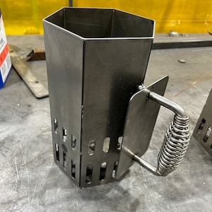 May include: A metal charcoal chimney starter with a hexagonal shape and a spring-loaded handle. The chimney starter has a series of holes on the sides for air circulation.