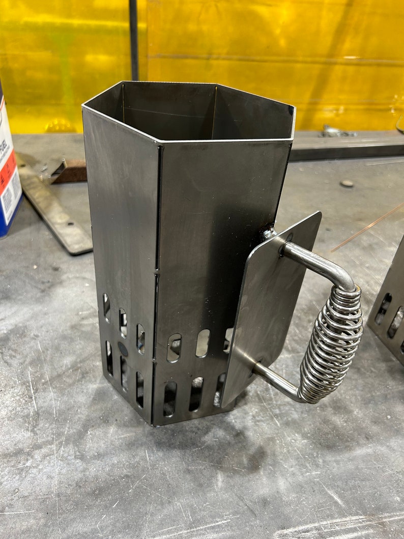 May include: A metal charcoal chimney starter with a hexagonal shape and a spring-loaded handle. The chimney starter has a series of holes on the sides for air circulation.