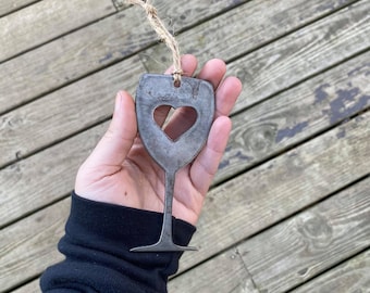 Puede incluir: Un adorno de copa de vino de metal con un corazón recortado. El adorno cuelga de una cuerda de yute.