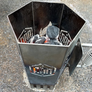 May include: A black metal charcoal chimney starter with a hexagonal shape and a handle. The chimney is filled with charcoal briquettes and has a small flame visible at the bottom.