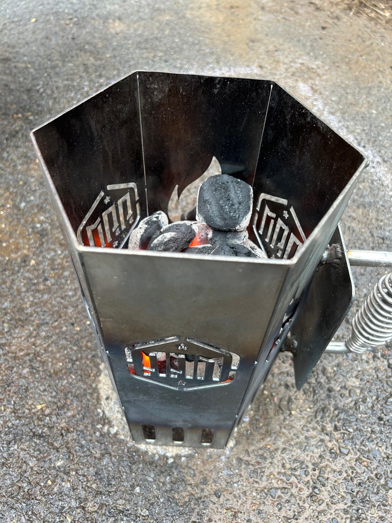 May include: A black metal charcoal chimney starter with a hexagonal shape and a handle. The chimney is filled with charcoal briquettes and has a small flame visible at the bottom.
