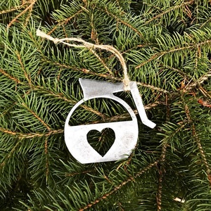 May include: A metal coffee pot ornament with a heart cutout, hanging on a green pine tree branch. The ornament is a rustic silver color.