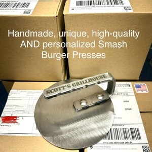 May include: A stainless steel smash burger press with a handle and the words "SCOTT'S GRILLHOUSE" engraved on the top. The press is sitting on top of a cardboard box with a shipping label.