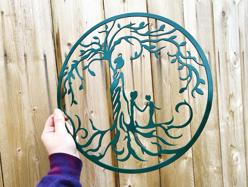Puede incluir: Una escultura de pared de metal azul verdoso que representa un &aacute;rbol de la vida con tres figuras de pie debajo de las ramas. El &aacute;rbol est&aacute; intrincadamente detallado con ramas y hojas que se arremolinan.