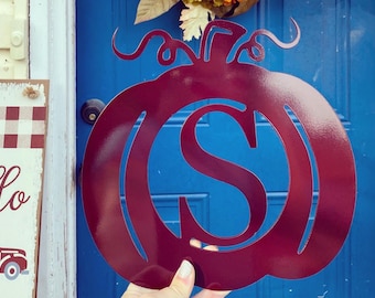 May include: A maroon metal pumpkin with a cut-out letter "S" in the center. The pumpkin is held in front of a blue door with a fall wreath.