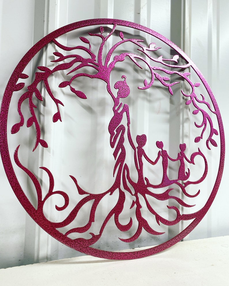 Puede incluir: Una pieza de arte de pared de metal rosa que representa un &aacute;rbol de la vida estilizado con una madre y tres hijos tomados de la mano. El &aacute;rbol tiene ramas y ra&iacute;ces intrincadas.