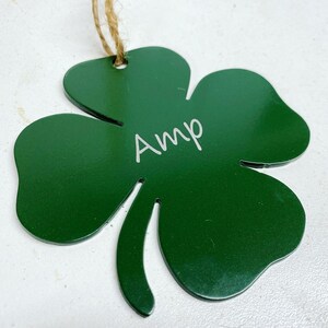 May include: A green metal shamrock ornament with the name "Amp" engraved on it. The ornament is attached to a twine string.