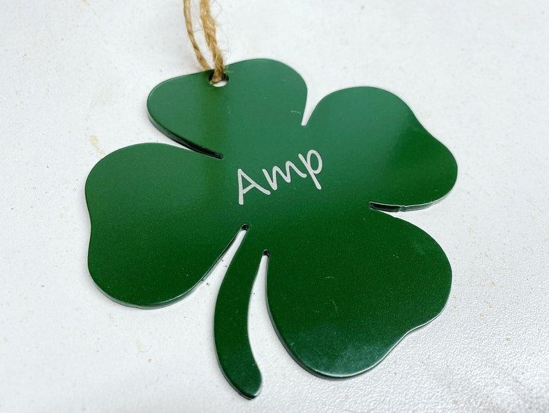 May include: A green metal shamrock ornament with the name "Amp" engraved on it. The ornament is attached to a twine string.
