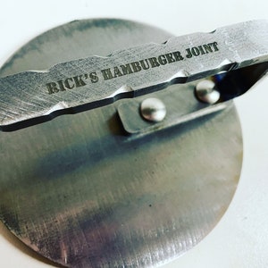 May include: A silver metal hamburger press with a handle that reads "RICK'S HAMBURGER JOINT".