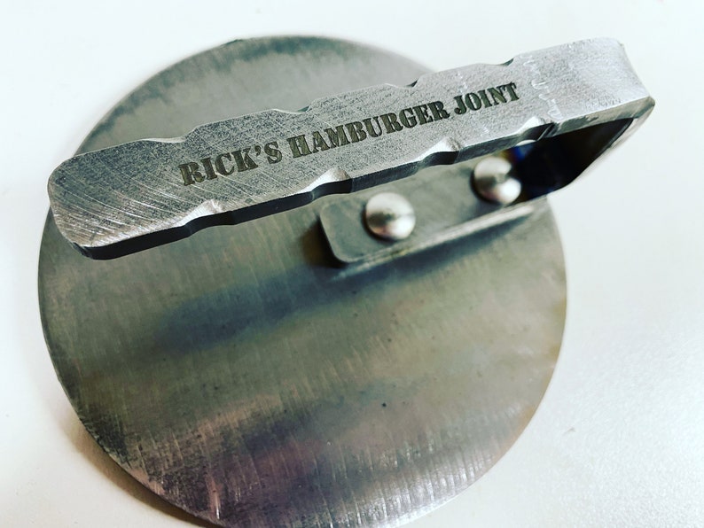 May include: A silver metal hamburger press with a handle that reads "RICK'S HAMBURGER JOINT".