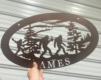 May include: A brown metal oval sign with a silhouette of three Bigfoot figures in a forest setting. The sign has the name "AMES" written in white lettering at the bottom.