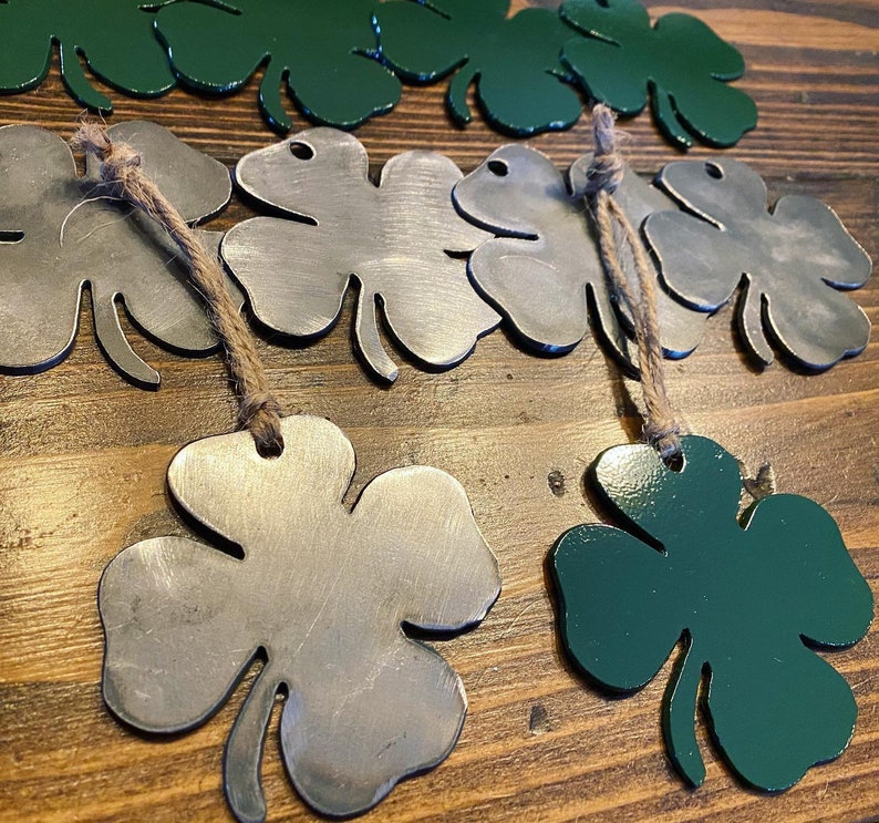 May include: A set of four-leaf clover ornaments, two are green and two are silver. The ornaments are made of metal and have a rustic look. They are hanging from twine.