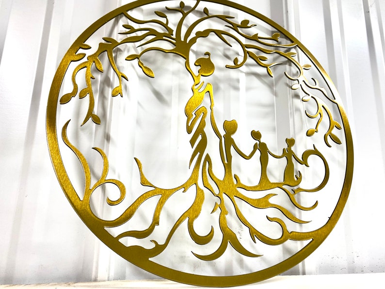 Puede incluir: Una decoraci&oacute;n de pared de metal dorado en forma de &aacute;rbol de la vida con una familia de tres figuras de pie sobre las ra&iacute;ces. El &aacute;rbol tiene ramas y hojas intrincadas.