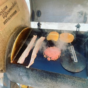May include: A close-up of a black metal grill with a metal lid that says "American Aftermarket Metalworks". The grill is open and there are two pieces of bacon, a hamburger patty, and two hamburger buns on the grill. There is steam coming from the grill.