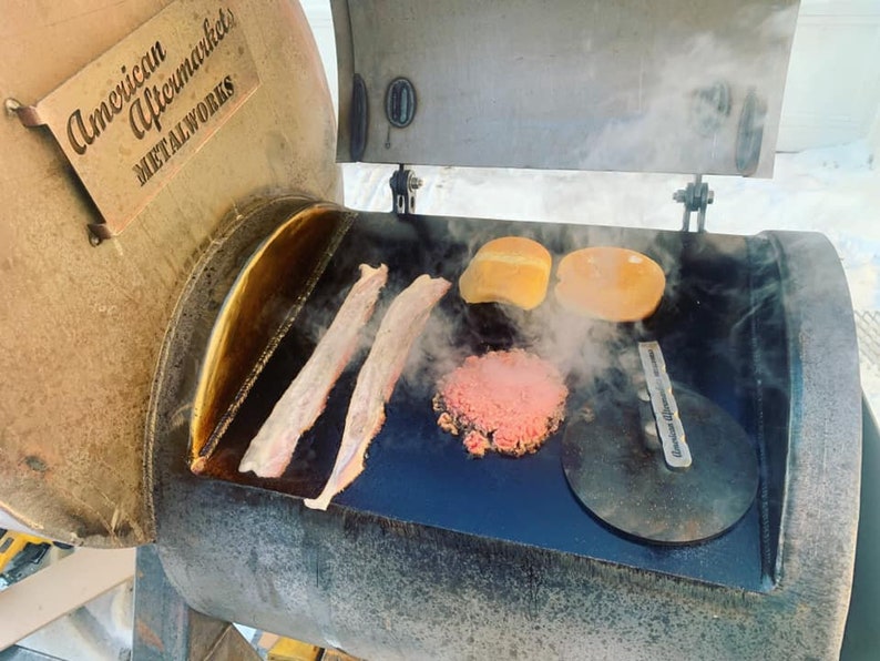 May include: A close-up of a black metal grill with a metal lid that says "American Aftermarket Metalworks". The grill is open and there are two pieces of bacon, a hamburger patty, and two hamburger buns on the grill. There is steam coming from the grill.