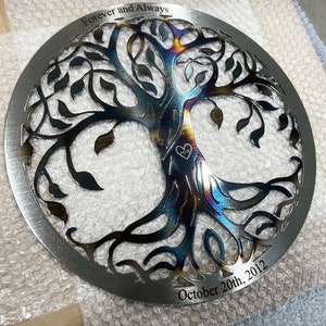 Peut inclure: Une &oelig;uvre d'art murale circulaire en m&eacute;tal repr&eacute;sentant un arbre de vie avec des branches et des feuilles complexes. Le m&eacute;tal a une finition arc-en-ciel iris&eacute;e et les mots "Forever and Always" et "October 20th, 2012" sont grav&eacute;s sur la pi&egrave;ce.