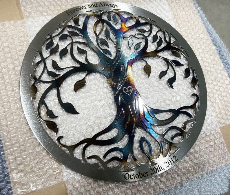 Peut inclure: Une &oelig;uvre d'art murale circulaire en m&eacute;tal repr&eacute;sentant un arbre de vie avec des branches et des feuilles complexes. Le m&eacute;tal a une finition arc-en-ciel iris&eacute;e et les mots "Forever and Always" et "October 20th, 2012" sont grav&eacute;s sur la pi&egrave;ce.