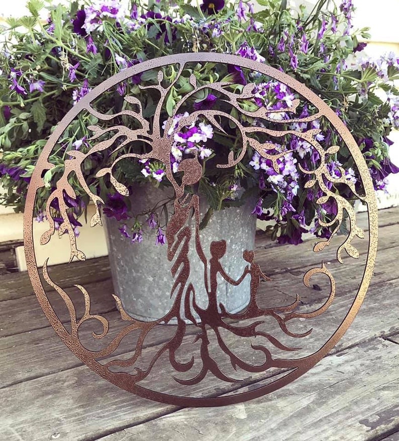 Puede incluir: Una decoraci&oacute;n de pared de metal marr&oacute;n en forma de &aacute;rbol de la vida con dos figuras de pie en la base del &aacute;rbol. El &aacute;rbol est&aacute; rodeado de flores moradas.
